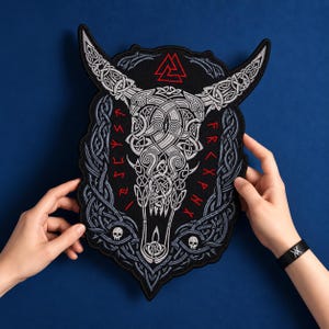 Könnte beinhalten: Ein großer gestickter Aufnäher mit einem weiß-grauen Stierschädel und komplexen keltischen Knotenmustern. Das Design enthält ein rotes Valknut-Symbol oben, rote Runenzeichen an den Seiten und zwei kleine Totenköpfe im unteren Bereich.