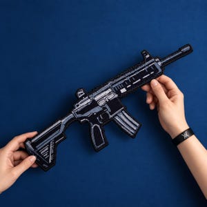 Könnte beinhalten: Ein schwarzer, gestickter Aufnäher in Form eines Gewehrs mit detaillierten weißen und hellblauen Nähten, die die Komponenten der Schusswaffe wie Lauf, Schaft, Magazin und Gehäuse vor einem einfarbigen dunkelblauen Hintergrund nachzeichnen.