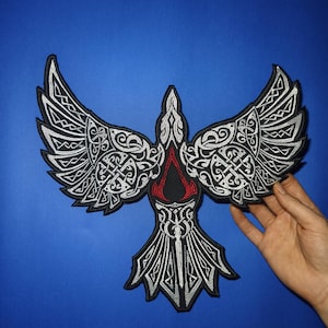 Könnte beinhalten: Ein gestickter Patch in Schwarzweiß, der einen stilisierten Vogel mit ausgebreiteten Flügeln zeigt. Der Vogel hat ein rotes und schwarzes Symbol auf der Brust. Das Symbol ist ein stilisierter Buchstabe "A" mit einer versteckten Klinge.