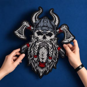 May include: An embroidered patch featuring a skull wearing a horned helmet with a long braided beard. Two crossed axes with intricate knotwork designs are positioned behind the skull. The embroidery uses black, white, and grey thread with red accents.