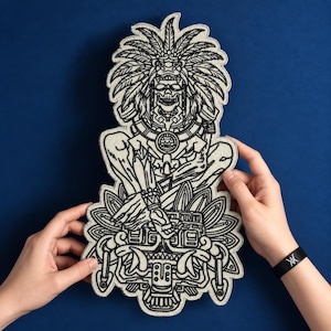 May include: A large embroidered patch featuring a detailed black and white illustration of a skeletal figure wearing an elaborate feathered headdress and ornate jewelry, seated above a complex decorative base with symmetrical patterns and lines.