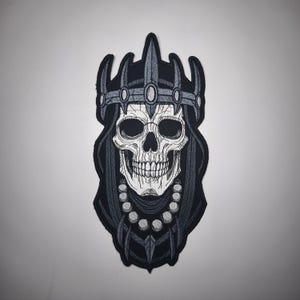 Puede incluir: Un parche negro y gris con una calavera que lleva una corona y una capa con capucha. La calavera tiene sombreado detallado y un collar de cuentas. El parche es para ropa o accesorios.