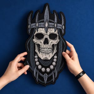 May include: An embroidered patch featuring a detailed skull wearing a spiked crown and a beaded necklace. The design uses black, white, and grey thread to create a high-contrast image of a skeletal figure with sharp, pointed crown elements.