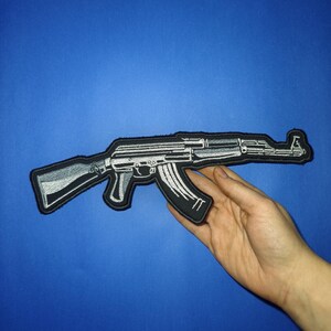 May include: A black and white embroidered patch depicting an AK-47 assault rifle. The patch is made of fabric and features a detailed design.