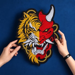 May include: A large embroidered patch featuring a split design of a yellow tiger face on the left and a red oni mask with a white horn on the right. The design includes intricate black stitching, white fangs, and a yellow rope border detail.