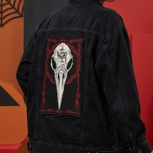 May include: A black denim jacket featuring a large rectangular patch on the back. The patch displays a white bird skull illustration with a black symbol on the forehead, framed by a red border with intricate red line patterns on a black background.