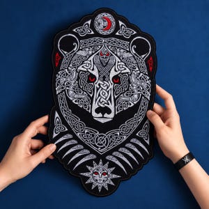 May include: A large black embroidered patch featuring a detailed bear head design with intricate white Celtic knotwork patterns. The bear has piercing red eyes, and the patch includes a red crescent moon symbol at the top and a sun motif at the bottom.