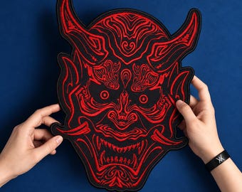 May include: A large embroidered patch featuring a red and black Hannya mask design. The patch displays intricate red thread detailing on a black fabric base, depicting a horned demon face with sharp teeth, wide eyes, and ornate facial patterns.