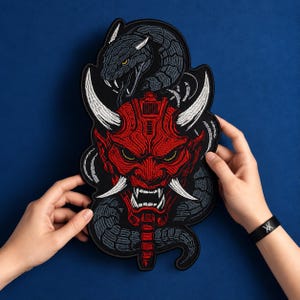 Peut inclure: Un grand écusson brodé représentant un masque oni rouge avec des cornes et des crocs blancs, entouré d'un serpent gris foncé enroulé aux yeux jaunes. Le motif présente des détails cousus complexes et une bordure noire sur fond bleu.