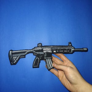 Puede incluir: Un parche bordado que representa un rifle de asalto negro y plateado con un cargador y una mira telescópica.