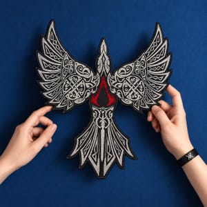Könnte beinhalten: Ein gestickter Aufnäher in Form eines Vogels mit ausgebreiteten Flügeln. Das Design zeigt komplizierte weiße und silberne keltische Knotenmuster auf schwarzem Grund, mit einem markanten roten Emblem in der Mitte des Brustbereichs.