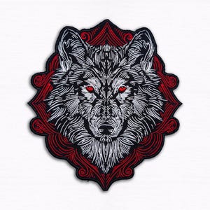 Peut inclure: Écusson brodé représentant une tête de loup aux yeux rouges perçants. Le loup est réalisé en fil blanc sur fond noir, encadré d'une bordure rouge décorative. Conçu pour être appliqué sur des vêtements ou accessoires.