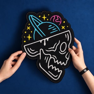Könnte beinhalten: Ein schwarzer bestickter Aufnäher mit einer weißen Totenkopf-Kontur und einem blauen Planeten mit Ringen darüber. Das Design enthält kleine gelbe Sterne und einen rosa Planeten auf einem dunklen Stoffuntergrund mit schwarzem Rand.