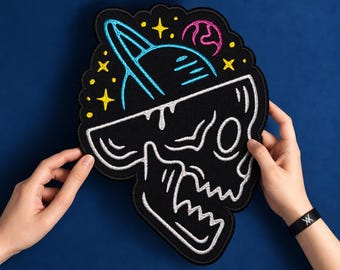 Op de afbeelding: Een zwarte geborduurde patch met een witte schedelomtrek en een blauwe planeet met ringen erbovenop. Het ontwerp bevat kleine gele sterren en een roze planeet op een donkere stoffen ondergrond met een zwarte rand.