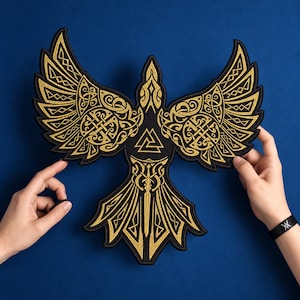 May include: A black embroidered patch shaped like a bird with wings spread wide. The design features intricate gold-colored knotwork patterns on the wings, a central triangular valknut symbol on the chest, and a sword motif on the tail feathers.