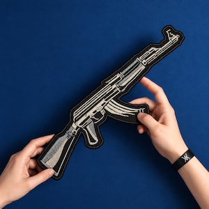 May include: An embroidered patch shaped like an AK-47 rifle, featuring a black background with detailed silver and grey thread work outlining the barrel, receiver, stock, and curved magazine, finished with a black stitched border.