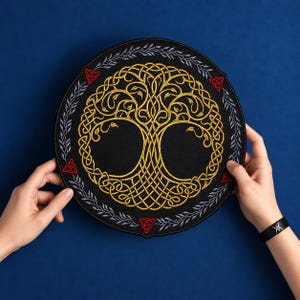 May include: A circular black embroidered patch featuring a central golden Celtic tree of life design. The border is decorated with silver leaf patterns and small red triquetra symbols. The embroidery is detailed with intricate knotwork.