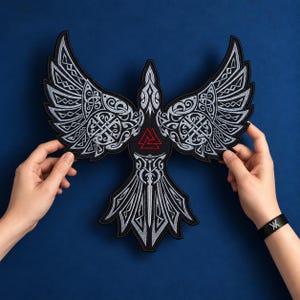 Peut inclure: Un grand écusson brodé représentant un corbeau noir stylisé aux ailes déployées. Le motif intègre des entrelacs celtiques complexes en gris argenté et un symbole Valknut rouge au centre de la poitrine, le tout sur un fond noir.