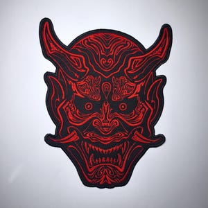 May include: An embroidered patch featuring a black background with a detailed red thread design of a horned mask. The mask displays intricate swirling patterns, pointed ears, and a wide mouth with visible teeth, all outlined in vibrant red stitching.