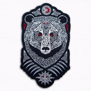 Op de afbeelding: Een zwart-wit geborduurde patch met een berenkop met ingewikkelde Keltische knoopwerk. De beer heeft rode ogen en een halve maan erboven. Onderaan staat een zon met een gezicht.