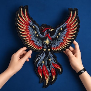 May include: A large embroidered patch featuring a phoenix with wings spread wide. The bird has black, red, and blue feathers with gold star accents and a prominent gold star on its chest. The design is detailed with intricate stitching throughout.
