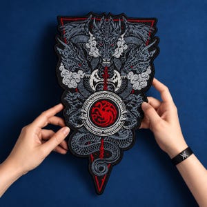 Könnte beinhalten: Ein großer, dreieckiger bestickter Aufnäher mit einem detaillierten dreiköpfigen Drachenmotiv in Grau- und Weißtönen mit roten Akzenten. In der Mitte befindet sich ein kreisförmiges Emblem mit einem roten dreiköpfigen Drachen auf schwarzem Grund.