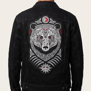 May include: A black denim jacket featuring a large embroidered back patch. The patch displays a white bear head with intricate Celtic knotwork, red eyes, and a red crescent moon symbol at the top. A sun motif is positioned at the bottom of the design.