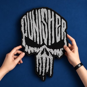 May include: A large, black, embroidered patch featuring the word PUNISHER in stylised, textured silver-grey thread. The lettering forms the shape of a skull, with the word integrated into the upper portion and vertical drips forming the jaw.