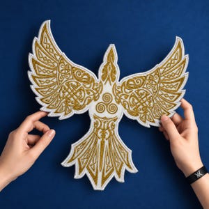 May include: An embroidered patch shaped like a bird with wings spread wide. The design features intricate gold metallic thread detailing on a white background, including Celtic-inspired knotwork patterns and a central triskelion symbol on the chest.