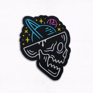 Può includere: Toppa a forma di teschio nero con un design a pianeta blu neon e stelle gialle. La toppa ha un contorno bianco e un pianeta rosa. La testa del teschio è aperta, rivelando la scena spaziale.