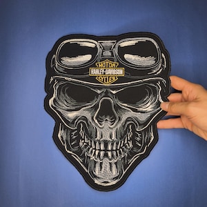 May include: Black and white embroidered patch featuring a skull wearing goggles. The patch has the Harley-Davidson Cycles logo in gold and white. The patch is held against a blue background.