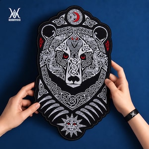Könnte beinhalten: Ein großer, schwarzer, bestickter Aufnäher mit einem detaillierten Bärenkopf-Design und komplizierten weißen keltischen Knotenmustern. Der Bär hat stechende rote Augen, und das Design umfasst eine rote Mondsichel oben und ein Sonnenmotiv unten.