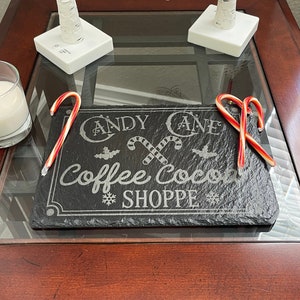 May include: A dark gray rectangular slate sign with the words "Candy Cane Coffee Cocoa Shoppe" etched in white. Two red and white candy canes and a glass of white liquid sit on a glass-topped table. Decorative birch tree branches are in the background.