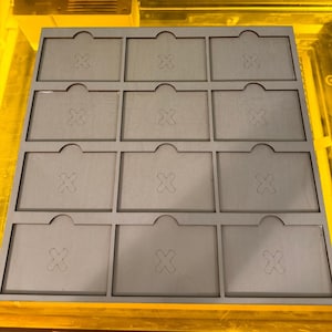May include: A grey wooden organiser with twelve rectangular compartments. Each compartment has a curved top and an 'X' cut out in the centre. The organiser is designed for small items and is likely used for storage or display.