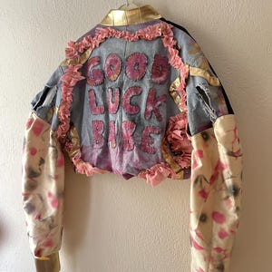 Può includere: Una giacca corta in denim con la scritta GOOD LUCK BABE in lettere rosa cucite sul retro. La giacca presenta colletto e polsini metallizzati dorati, rifiniture con volant rosa e maniche lunghe con un motivo tie-dye color crema, rosa e grigio.