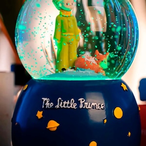 Op de afbeelding: Een sneeuwbol met een scène uit "De Kleine Prins". De blauwe basis is versierd met gele sterren en planeten. Binnenin zijn de Kleine Prins en een vos te zien. De bol is gevuld met sprankelende glitter.