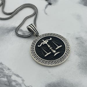 May include: A silver-colored pendant necklace featuring a round pendant with a detailed design. The pendant has a black background with a silver-colored emblem and a Greek key border. The chain is also silver-colored.