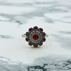 May include: A silver ring with a red and white gemstone surrounded by purple stones. The ring has a floral design.