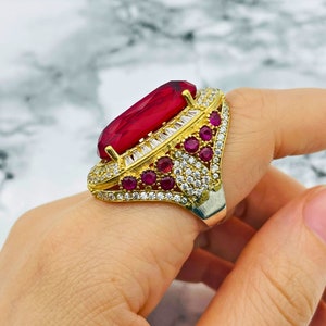 Peut inclure: Une bague en or avec une grande pierre rouge entourée de petites pierres rouges et de zircons cubiques blancs.
