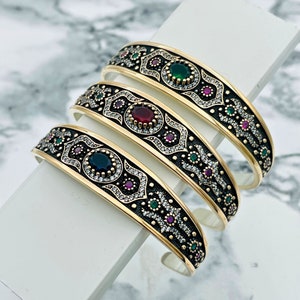 May include: Three gold-toned bracelets with intricate designs featuring black enamel, white stones, and colorful gemstones. The bracelets are stacked on top of each other, showcasing the details of each piece.