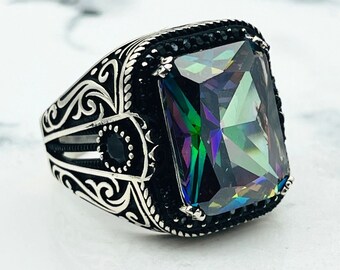May include: A silver ring with an ornate design and a large, faceted, iridescent gemstone. The gemstone is set in a square shape and surrounded by smaller black stones.