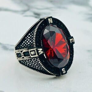 May include: A silver ring with a large red gemstone and black accents. The ring features a compass design with the letters N, E, S, and W around the gemstone.