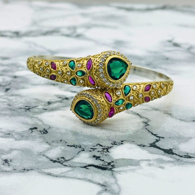 May include: A gold bangle bracelet with a snake design. The bracelet is adorned with green and pink gemstones and white cubic zirconia.