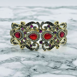 May include: Gold-tone bracelet with intricate filigree design and red and clear gemstones.