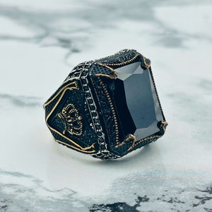 Peut inclure: Une bague noire et dorée avec une grande pierre rectangulaire noire. La bague a un design complexe et orné.