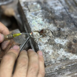 May include: A close-up of a jeweler's hands working on a silver ring with a red gemstone. The jeweler is using a small torch to heat the ring.