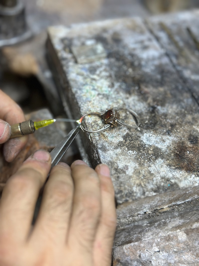 May include: A close-up of a jeweler's hands working on a silver ring with a red gemstone. The jeweler is using a small torch to heat the ring.