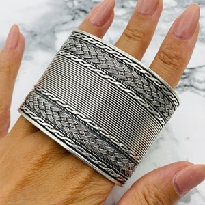 May include: A wide silver cuff bracelet with intricate braided and ribbed detailing. The bracelet is shown on a hand, showcasing the textured design and metallic sheen. A statement piece of jewelry.