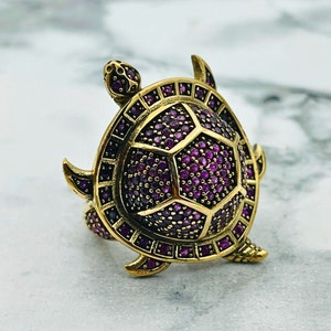 Puede incluir: Un anillo de tortuga dorado con una concha de piedras preciosas púrpuras. El anillo está detallado con intrincados tallados y un acabado texturizado.