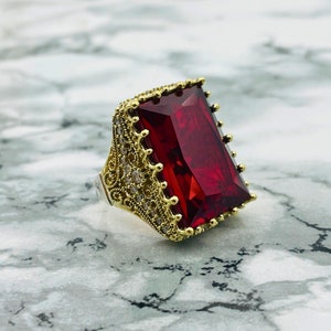 May include: A gold ring with a large, rectangular, red gemstone set in a filigree design. The ring is surrounded by smaller, clear gemstones.